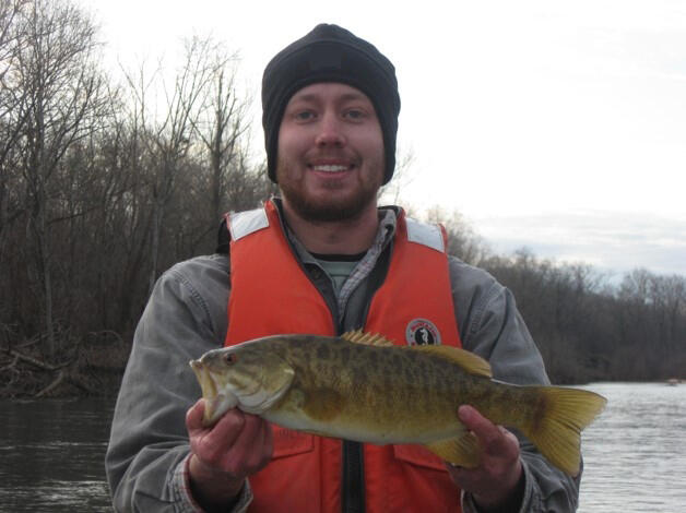 male scientist, smiling with a beard, wearing an orange personal floatation device, gray sweatshirt, black hat, standing near water