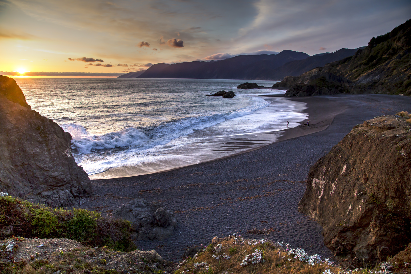 Waves crash on a rocky beach at sunset under a multicolored sky