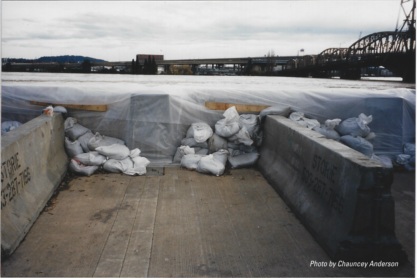 flood waters visible just below the wall supported by concrete blocks and sandbags