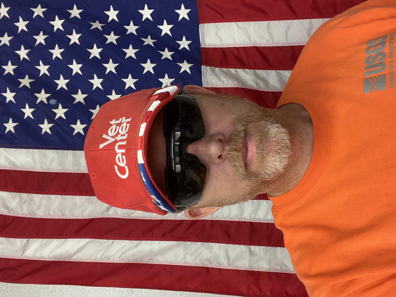 Man with orange USGS shirt, sunglasses, and red cap standing in front of an American flag.