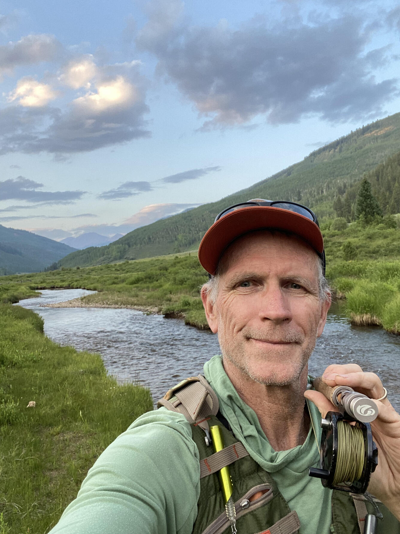 male scientist, smiling, holding a fly fishing rod, wearing a hat and green sweatshirt, standing near a river and mountains