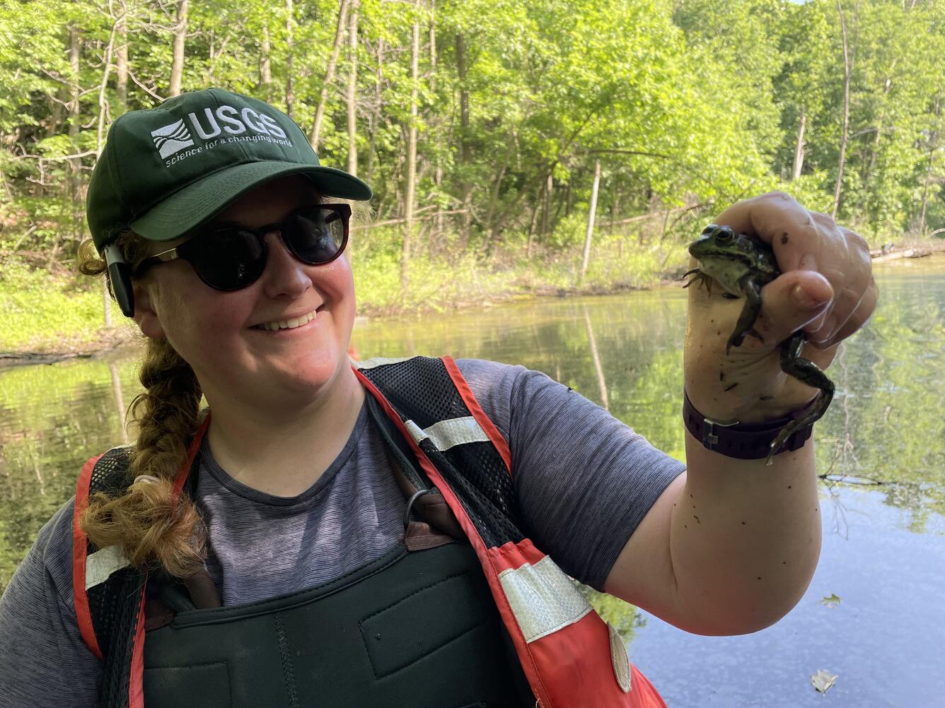 Megan standing in front of water and trees, holding a frog in one hand, and wearing a baseball cap, sunglasses, reflective vest, and overalls.