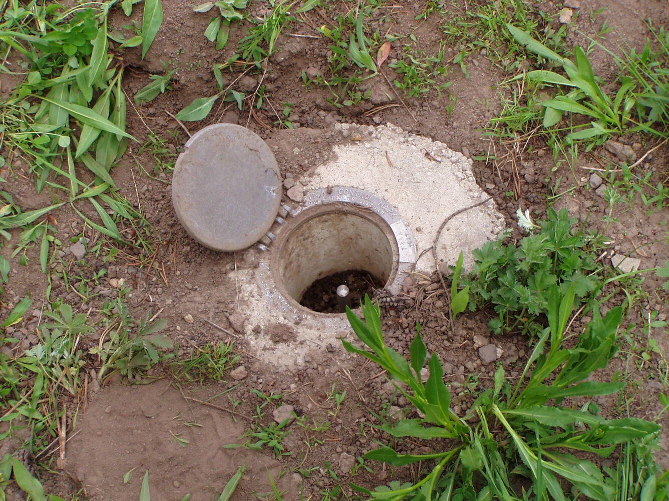 A hinged metal cover, flush with the ground, is open, exposing a metal rod just below the ground surface.