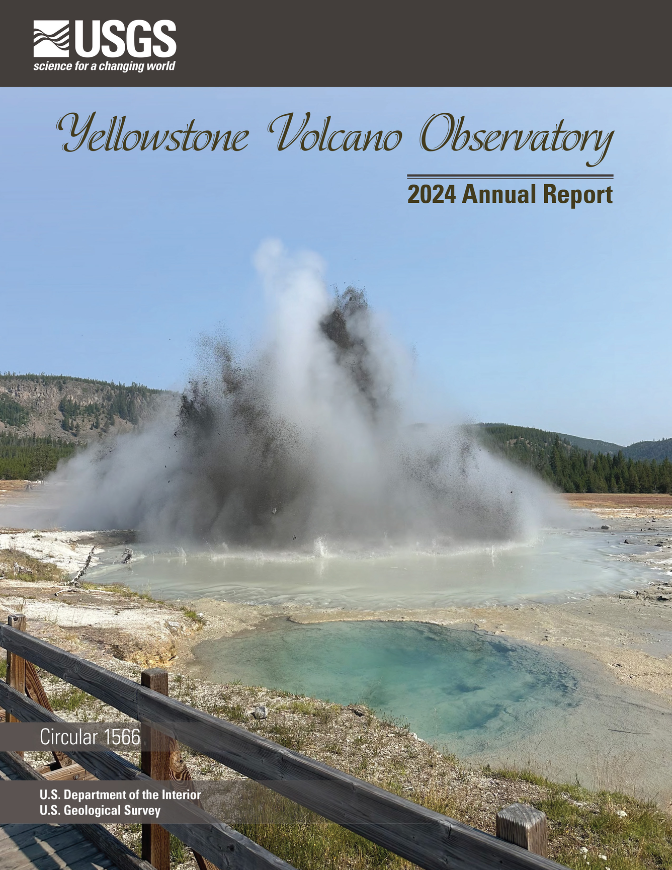 Muddy eruption from a blue water spring on barren ground, forested hills and blue sky in the background