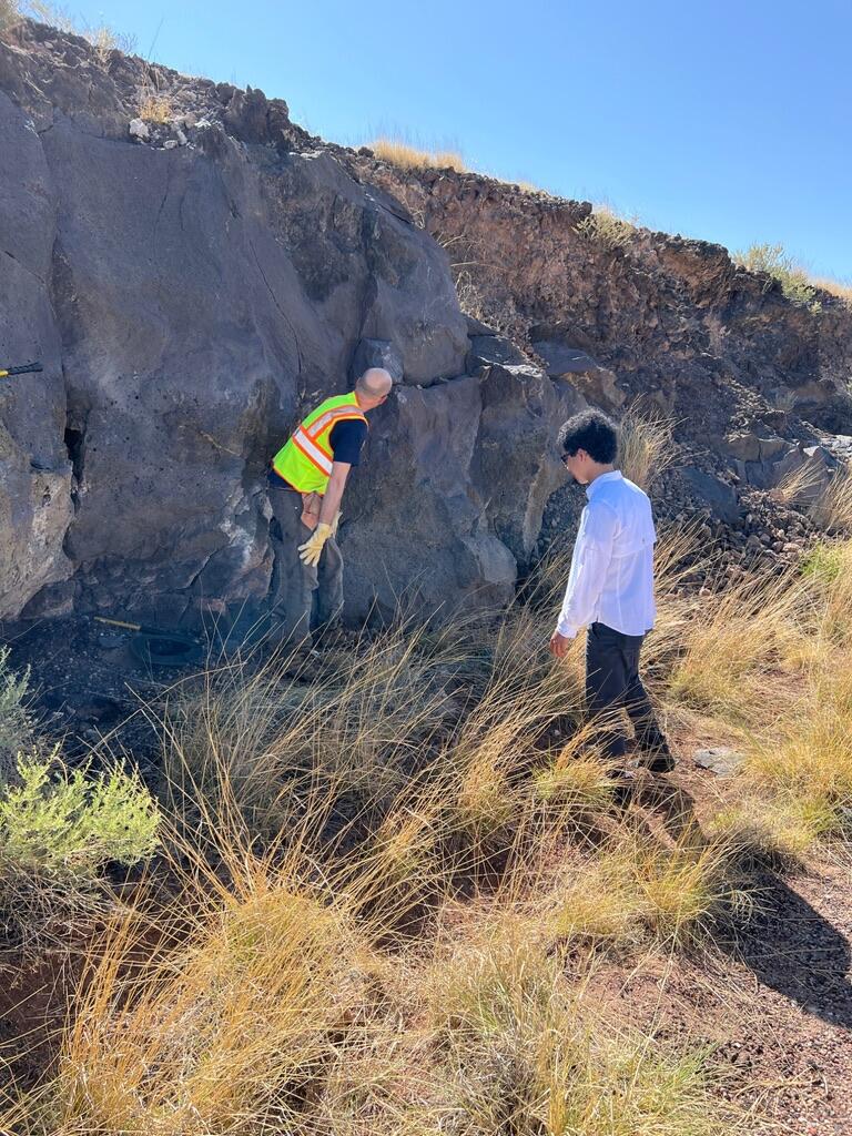 Two men examine a vertical outcrop of gray rock about twice their height, with brown grass at their feet and blue sky above