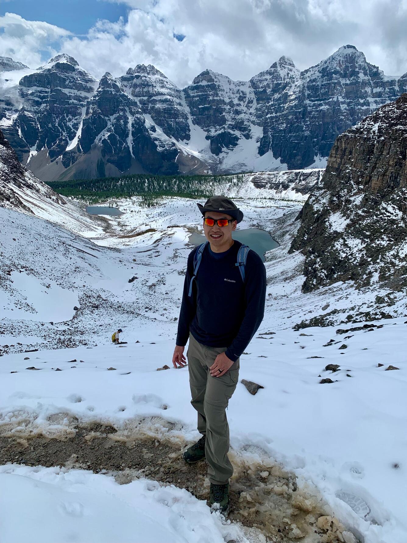 Ian Yuh is facing the camera wearing a gray brimmed hat, orange sunglasses, dark blue sweatshirt and khaki pants. The background is a snowy valley, with mountains and trees in the middle.