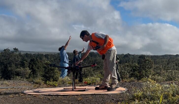 Color photo of a USGS scientist in an orange vest sitting up a UAS while two scientists in the background observe for birds