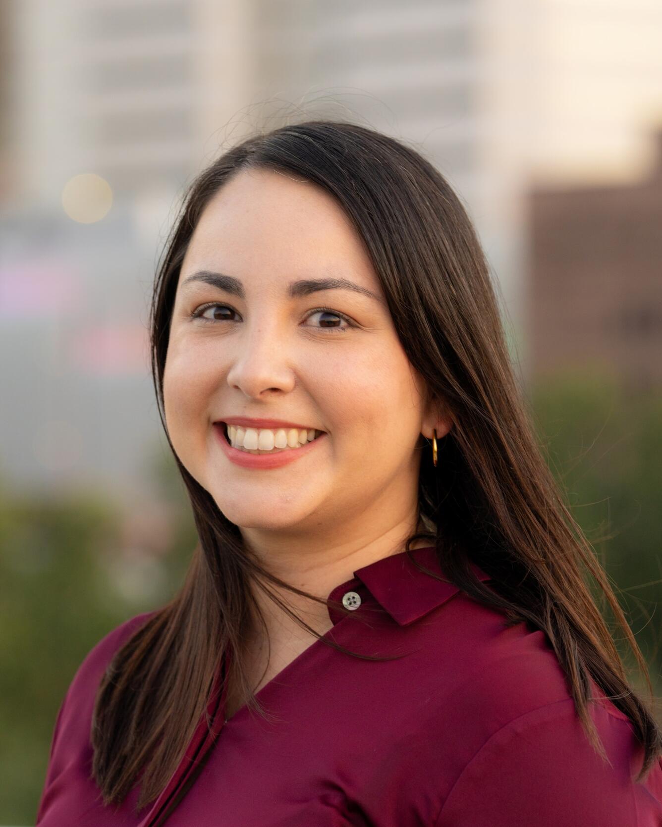 Color photo of Zulimar smiling. She has long brown straight hair and is wearing a marroon shirt with city buildings in the background. 