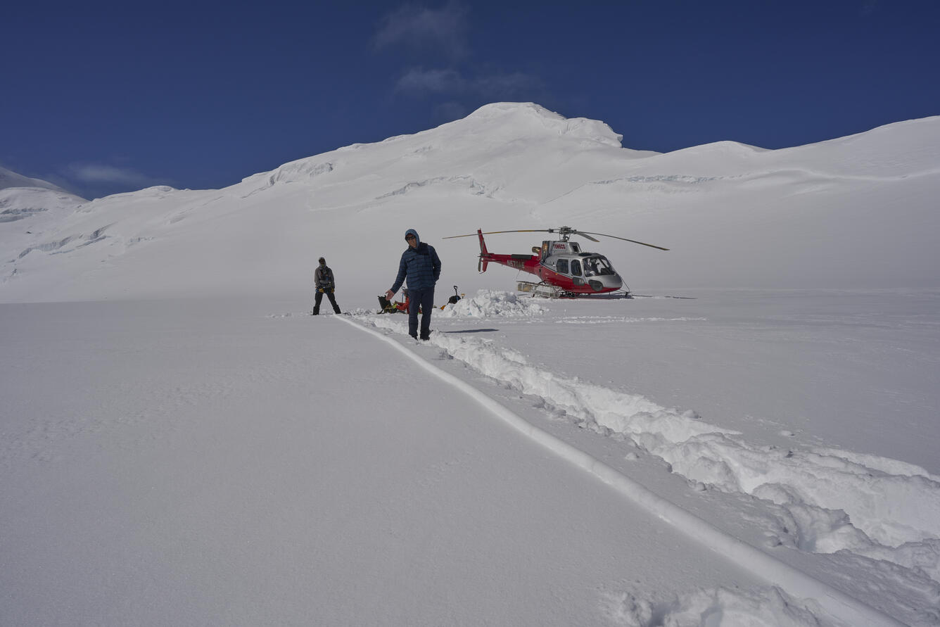 Two people standing in the snow next to a helicopter on top of a mountain.