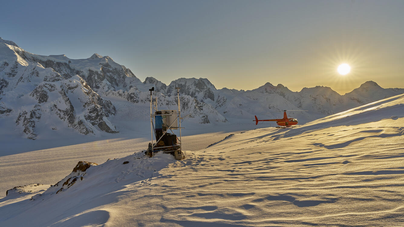 Person in blue jacket at a weather station on snow with the sunshine, mountains, and a red helicopter in the background