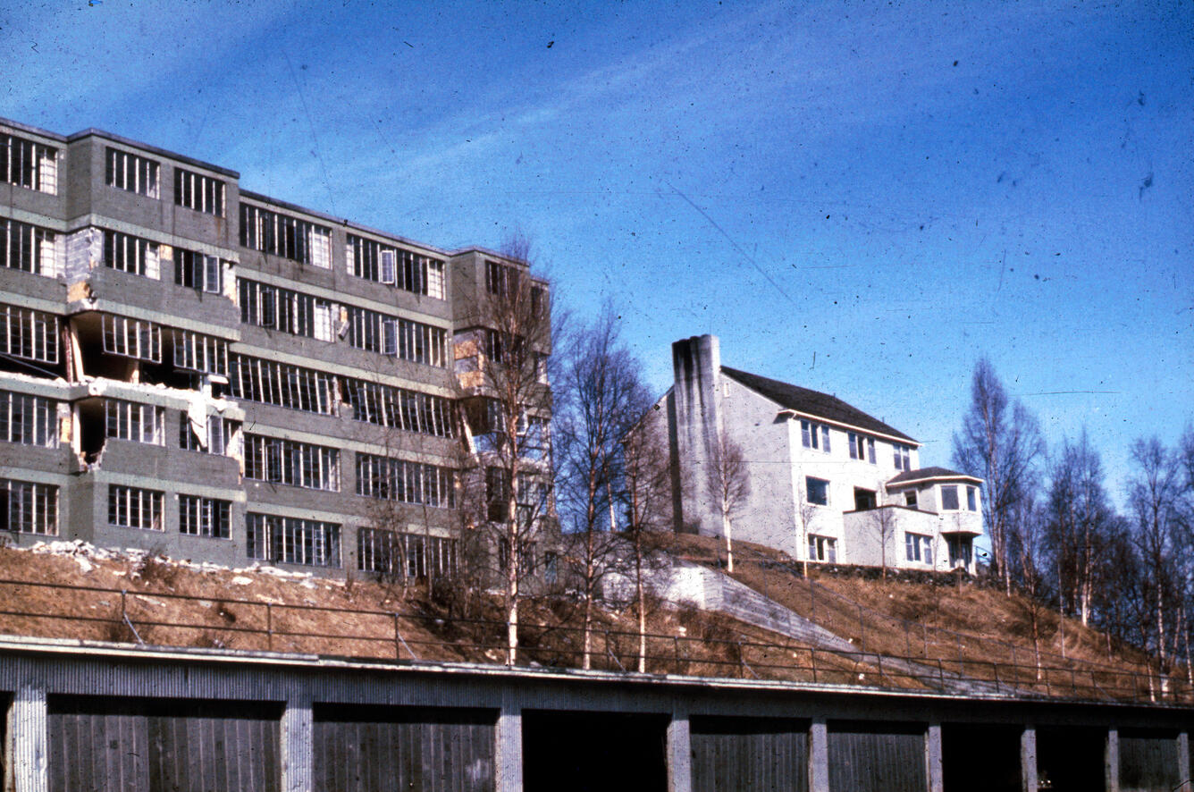 Two buildings on hill. One is damaged, exposed brick, no windows and other house has no damage. 