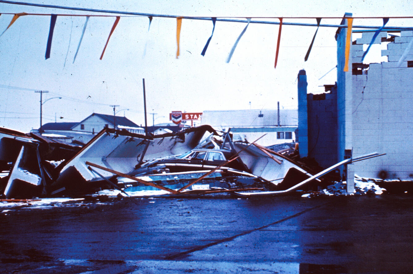 Building collapse. Roof debris crumbled on ground, colored flags hanging in foreground with Chevron gas station sign in back.