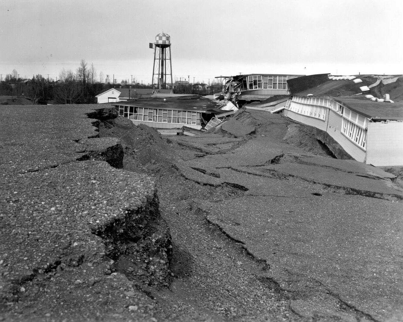 Ground that has shifted downward next to damages school. Part of building is engulfed in open ground. Water tower in back.