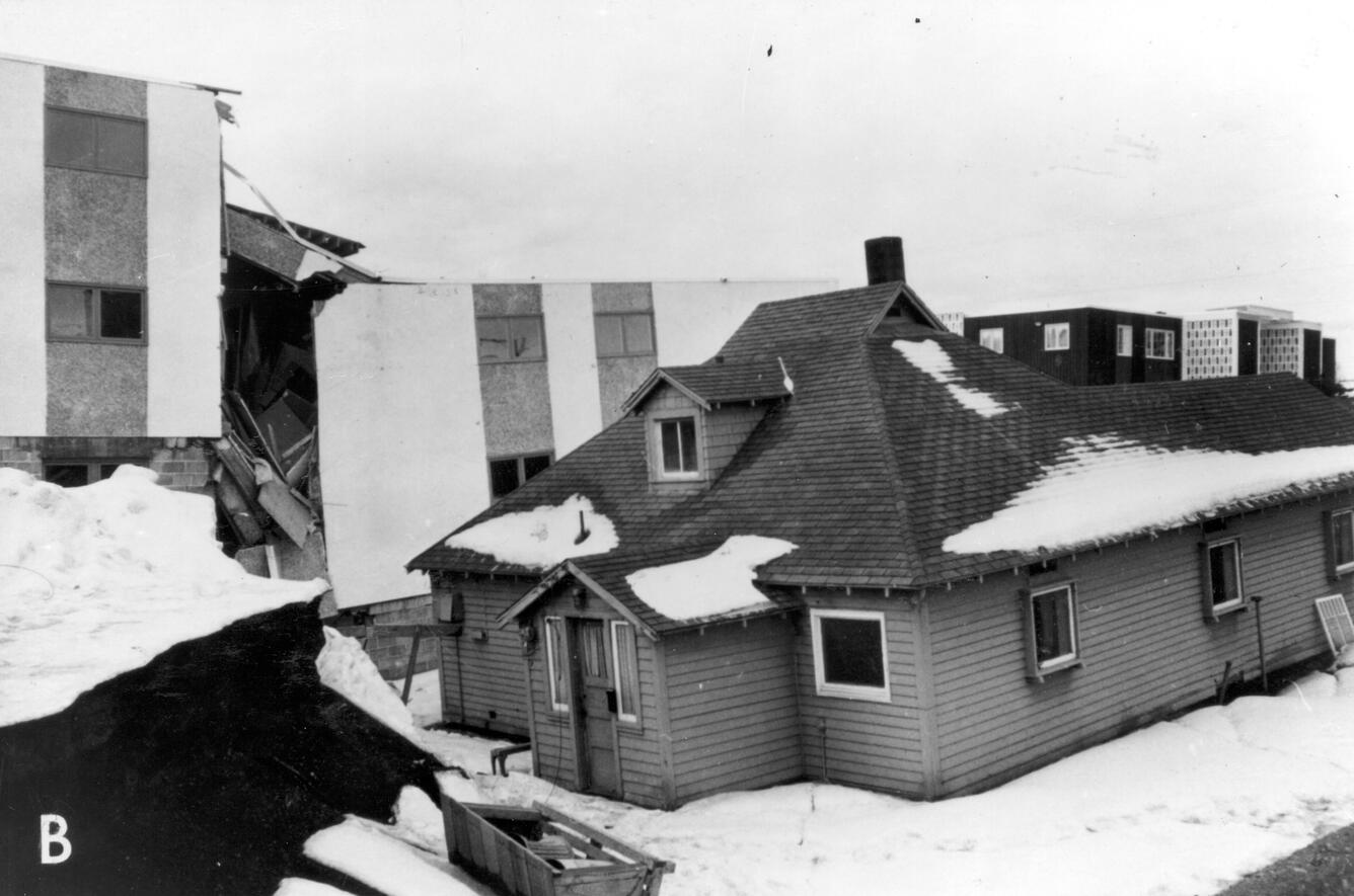 House surround by snow is undamaged and intact. Apartment building behind house is crumbling and broken in two. 