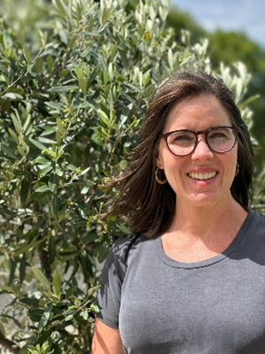 A woman with brown hair and a grey shirt stands in front of a thicket of trees.