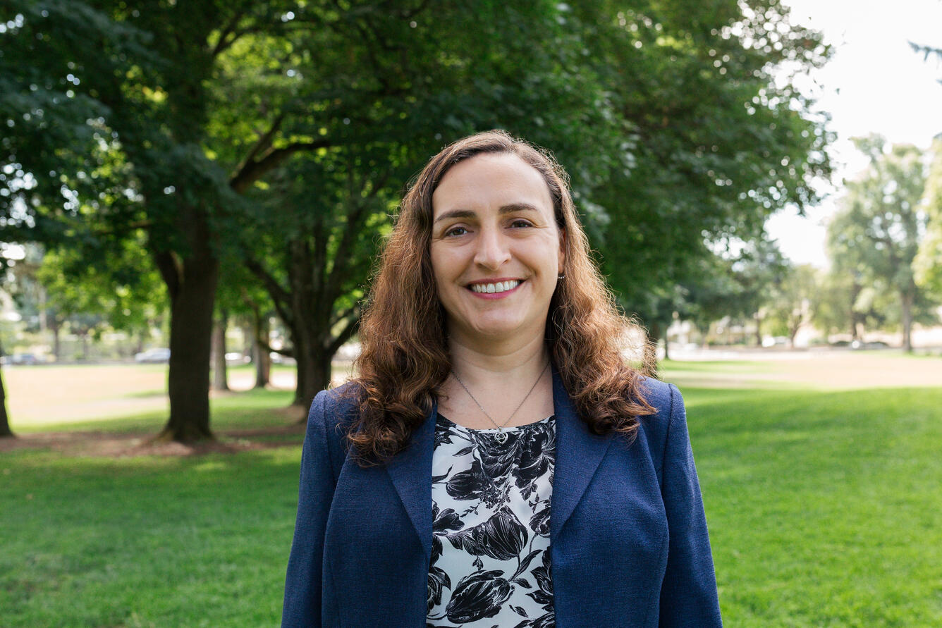 A woman with long dark hair and a black and white shirt with a navy jacket smiles for her picture while standing in a park.