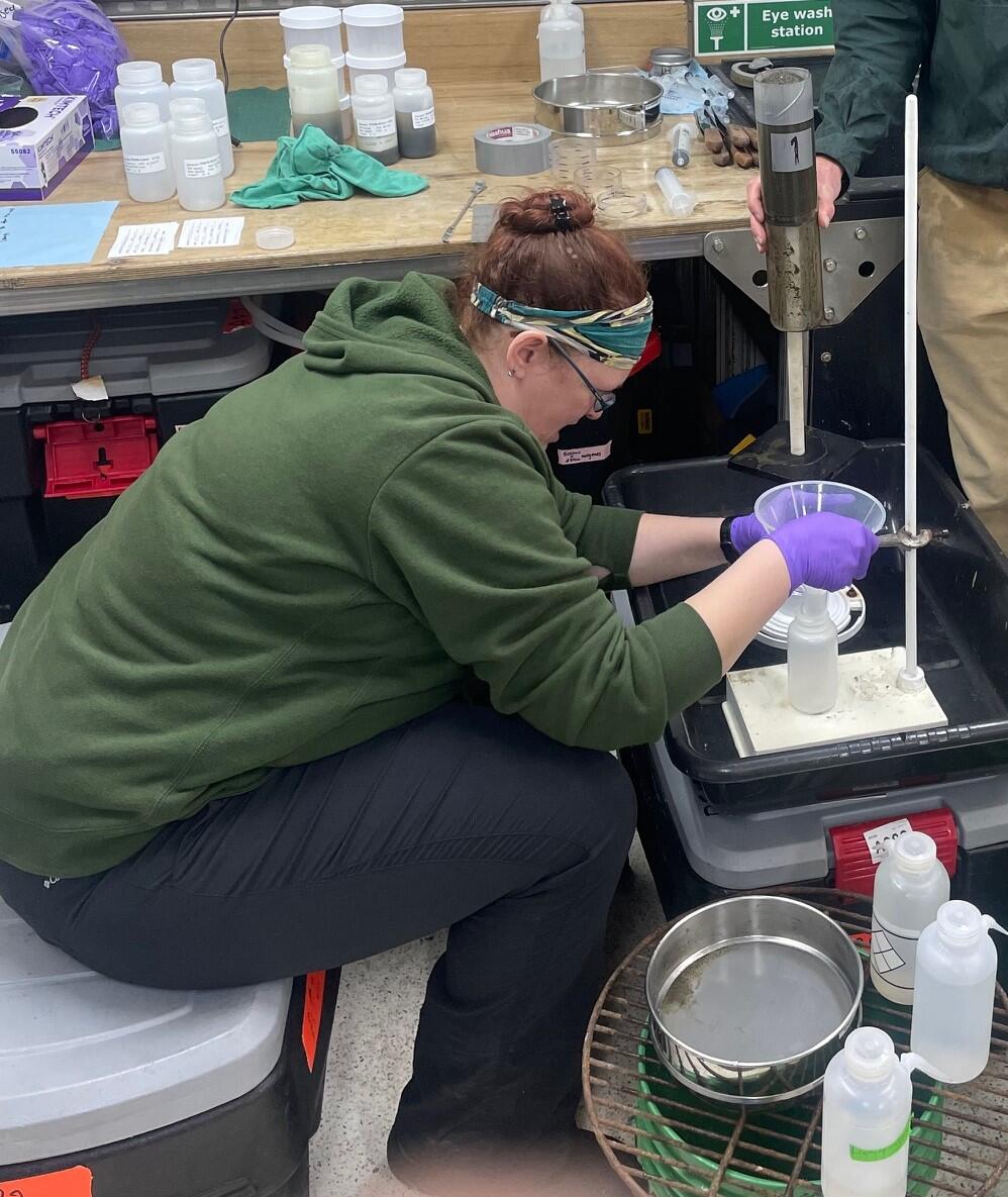 A person in a green hoodie and black pants sits on an action packer and holds a funnel as they process sediment