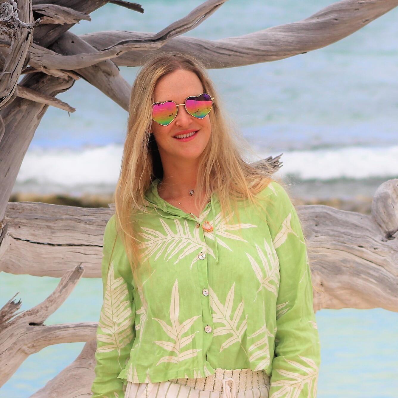 A blonde woman wearing a green patterned shirt and heart sunglasses stands next to driftwood on a beach