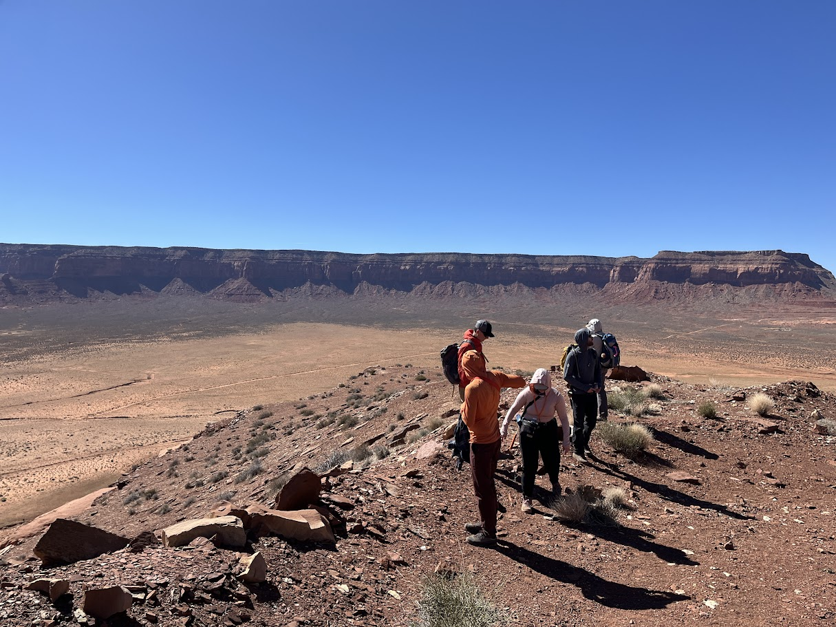 Five people stand in the Utah desert viewing the rocks around them with a large cliff in the background.