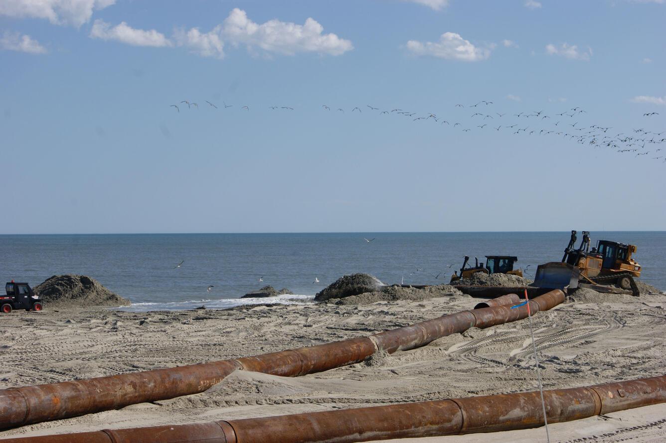 Rusty pipes lie on sandy beach with heavy equipment in the background moving the sand.