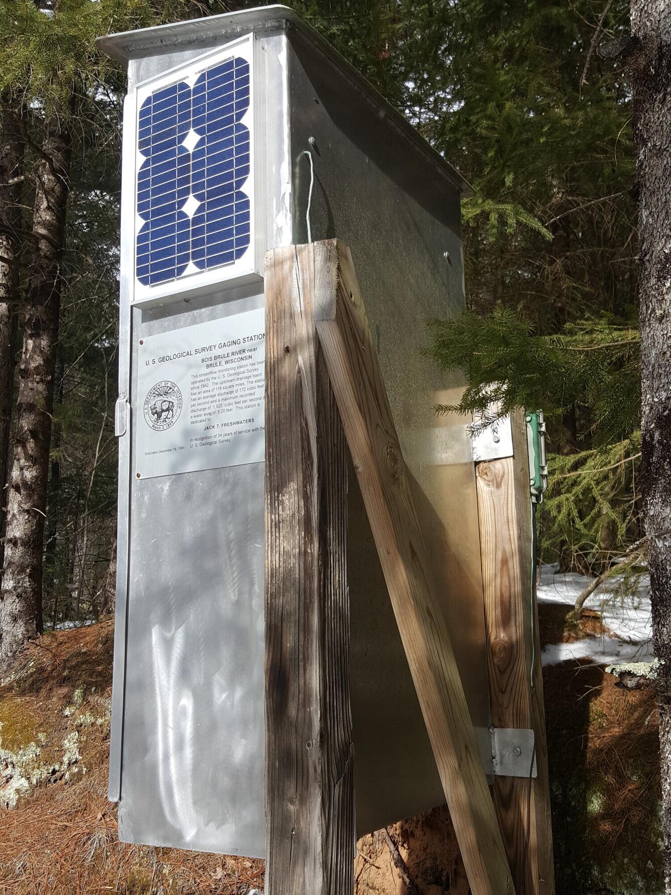 Gray metal USGS streamgage housing with solar panel and inscription mounted on a wooden support in a forested area.