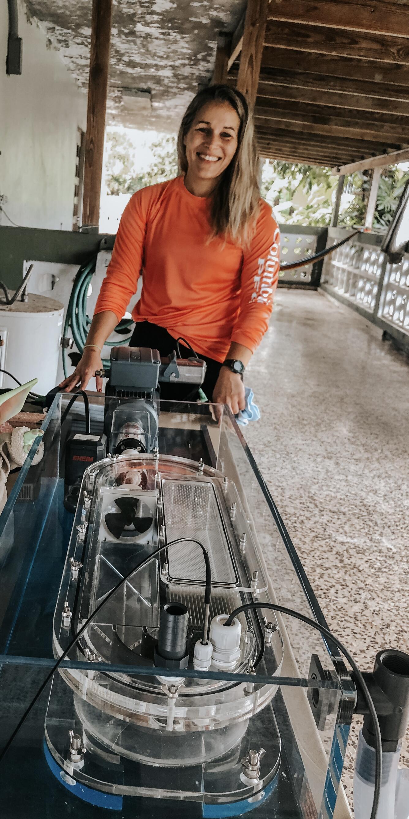 A woman with long blonde hair and wearing a long sleeved orange shirt standing in front of research equipment.