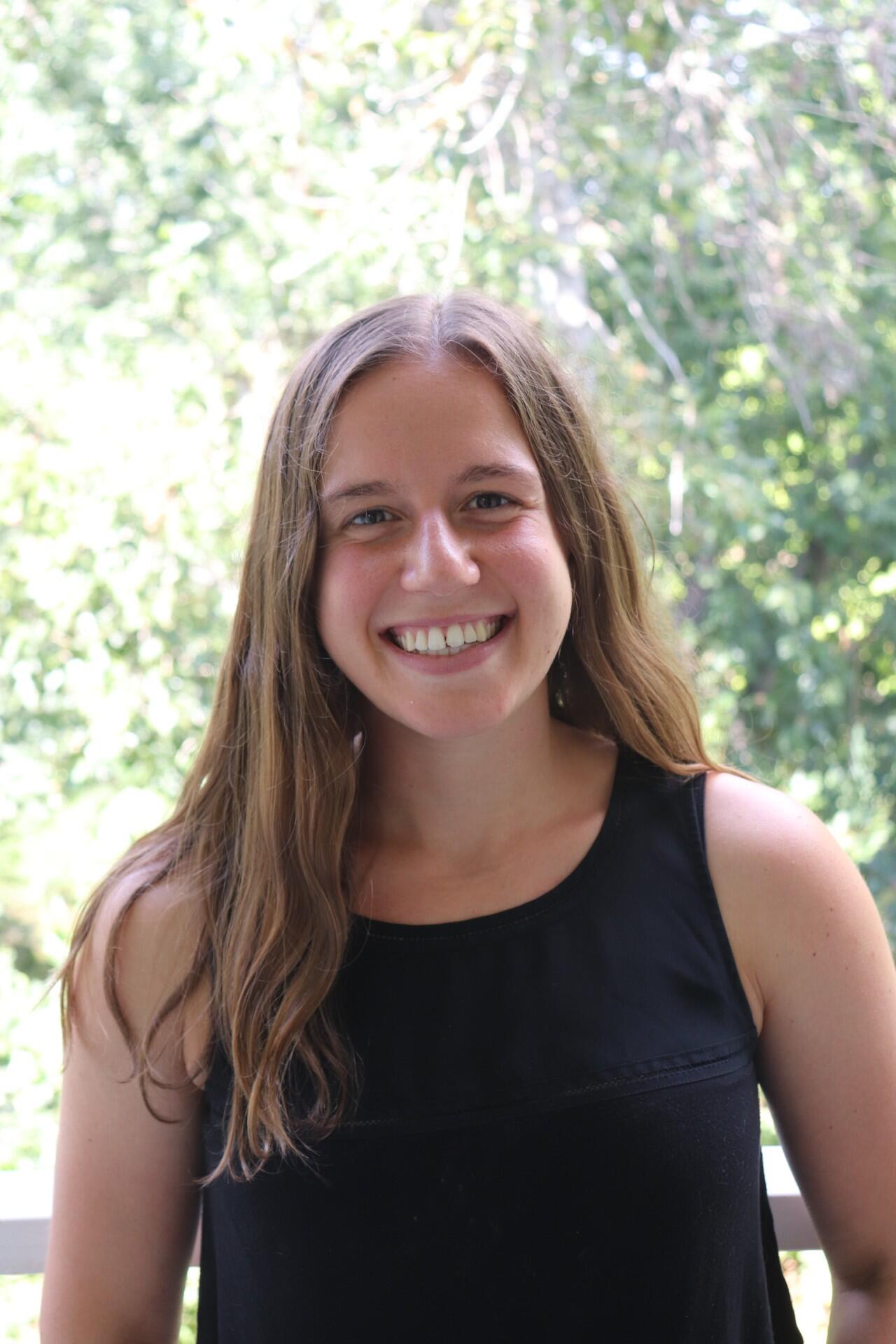Smiling woman with long brown hair standing against leafy green background.