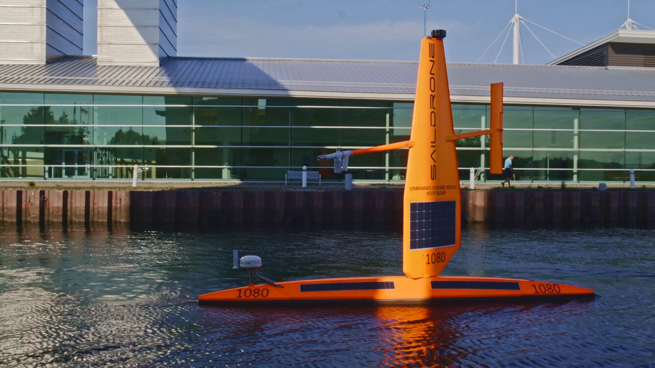 A Saildrone, a long skinny orange surface vehicle with a sail-like structure sticking out of it, in the harbor on Lake Erie