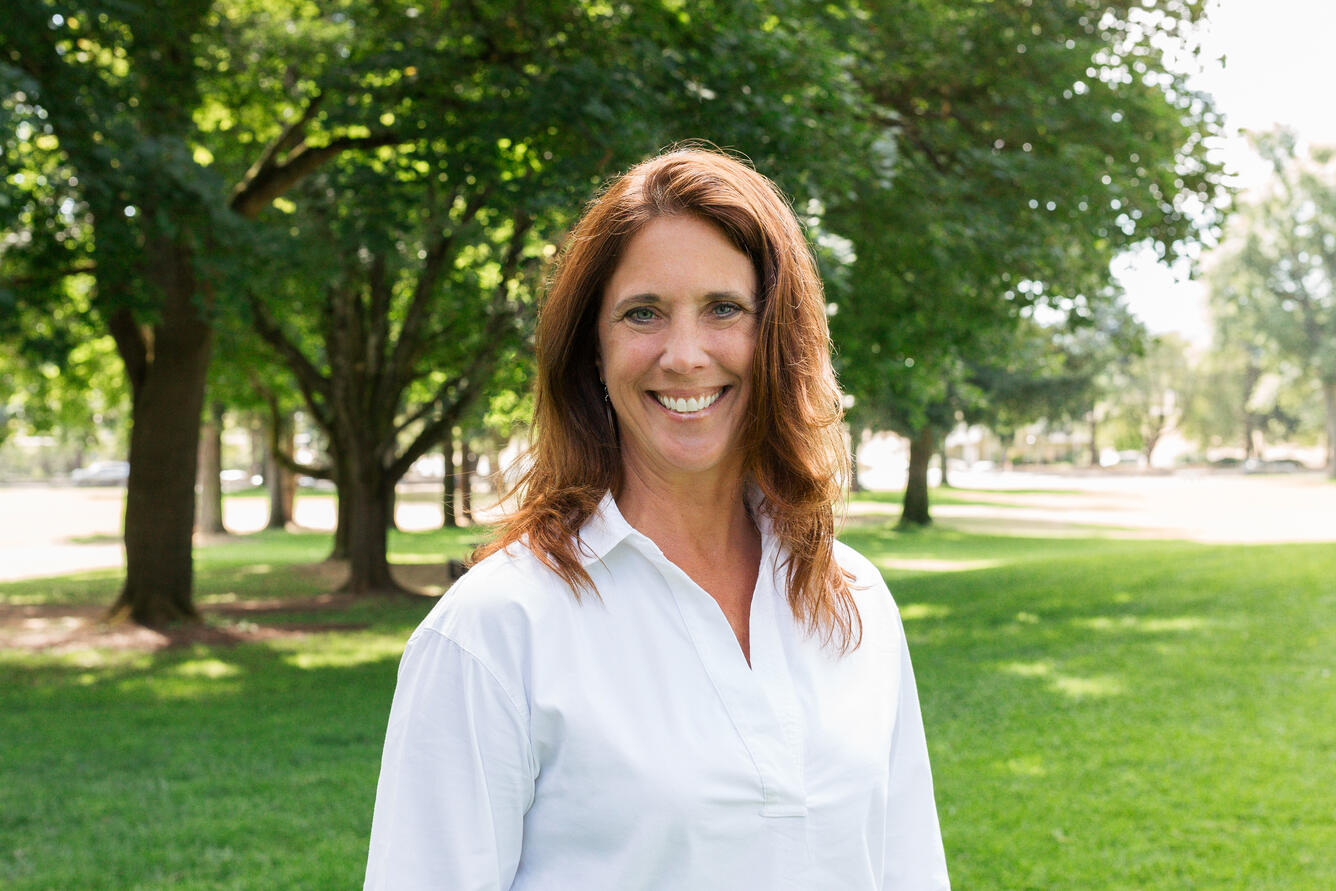 A woman with long brown hair is wearing a white shirt and smiling for her picture while standing in a park.