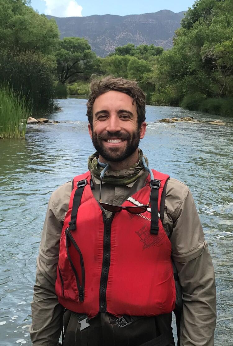 man with a red vest, brown hair, tan jacket, brown pants, standing near a river with trees and mountains