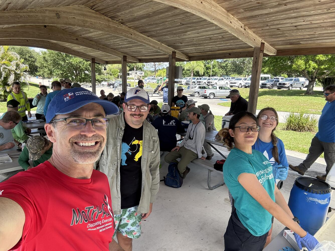 Four people stand in the foreground taking a selfie-style photo, with many people sitting on metal park benches under a large pavilion in the mid-ground, and a road and parking lot in the background