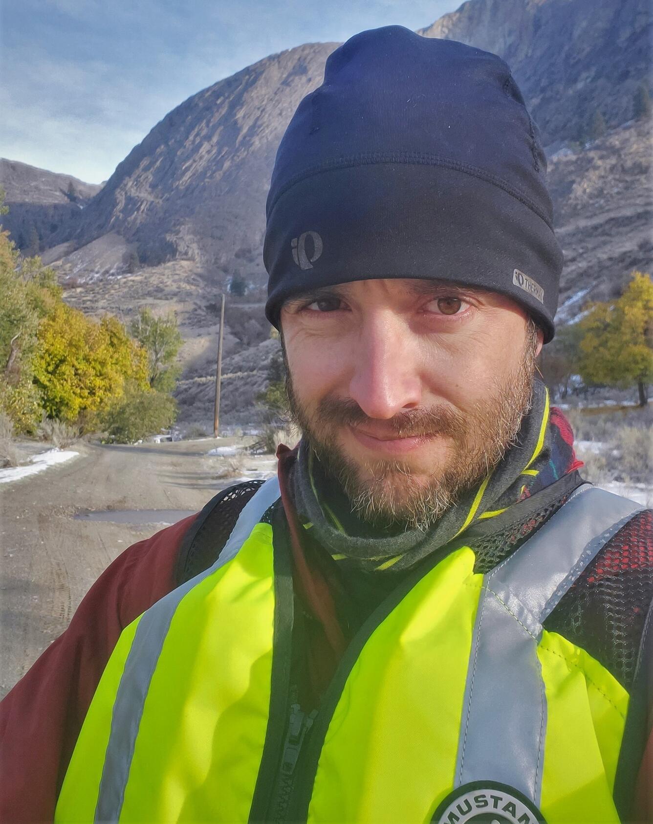 Headshot of man wearing warm hat and life vest in front of mountains