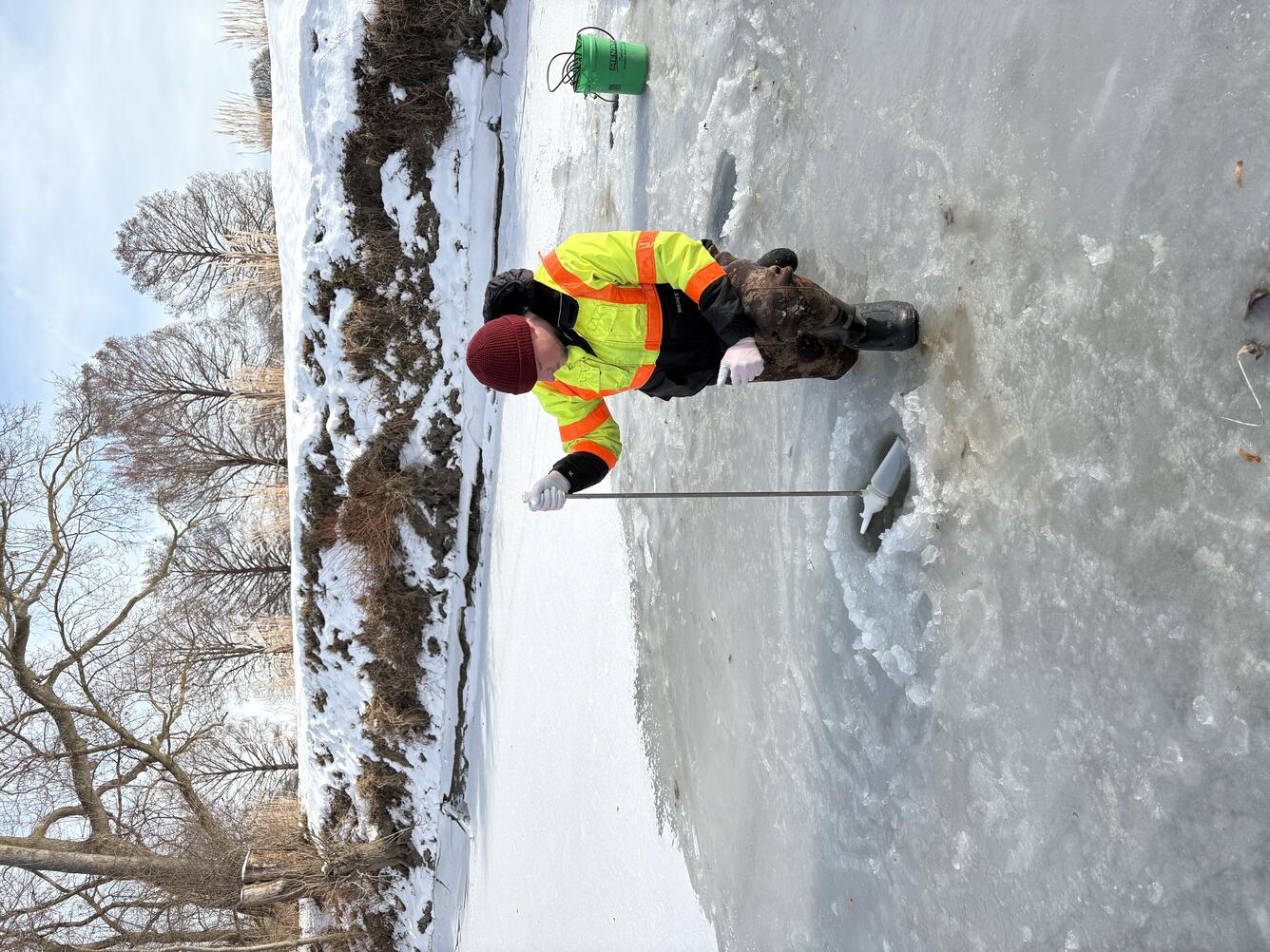 Hydrologic technician in bright yellow coat collects water sample through hole in ice on frozen river