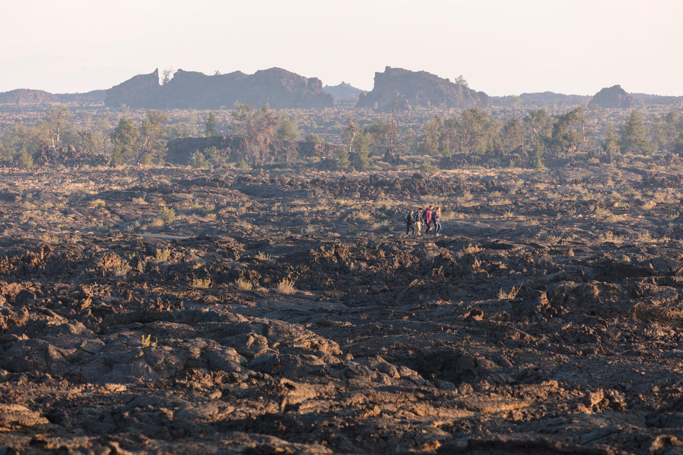 four people walk across a lava bed, trees and rocky hills in the background