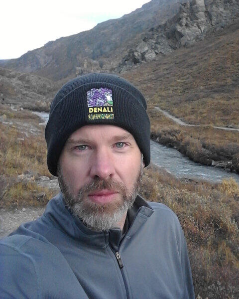 graduate student wearing a black hat, gray top, standing near water and mountains