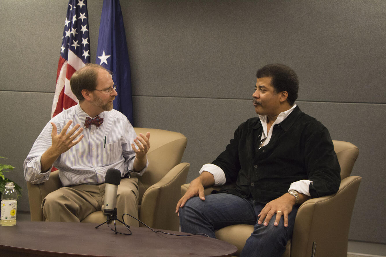 Image shows two men sitting in chairs with a table between them