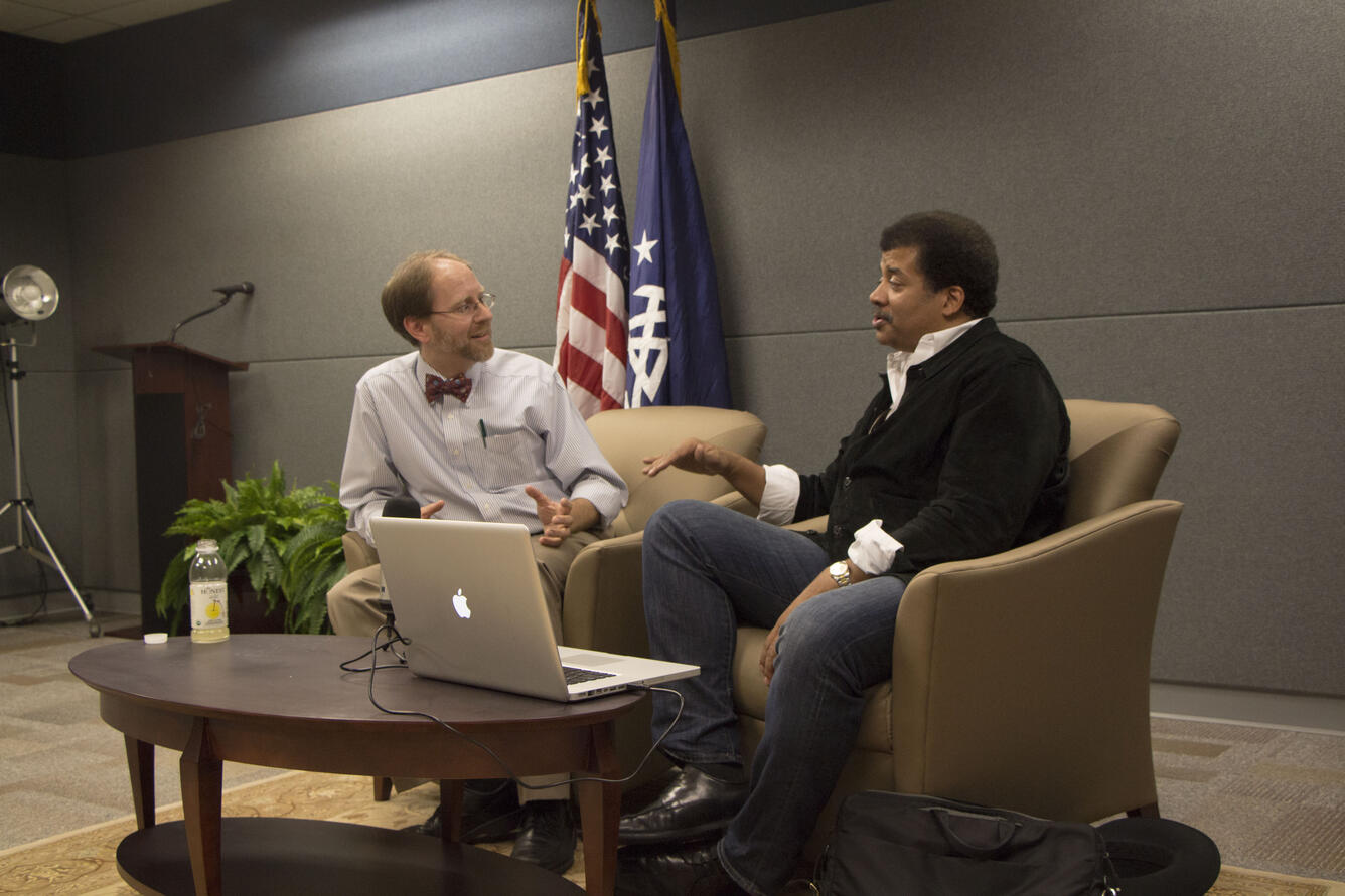 Image shows two men sitting in chairs with a table between them