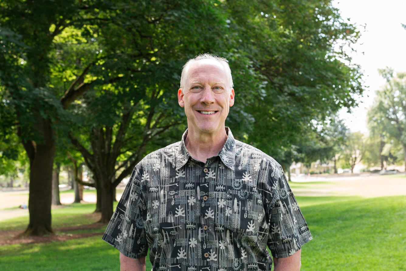 A man with short, grey hair wearing a black and white button up smiles for his picture in a park. 