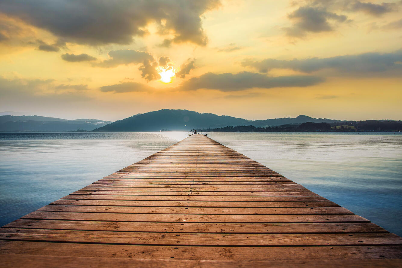 Image of dock looking out over water with sun and clouds in the sky and land in distance
