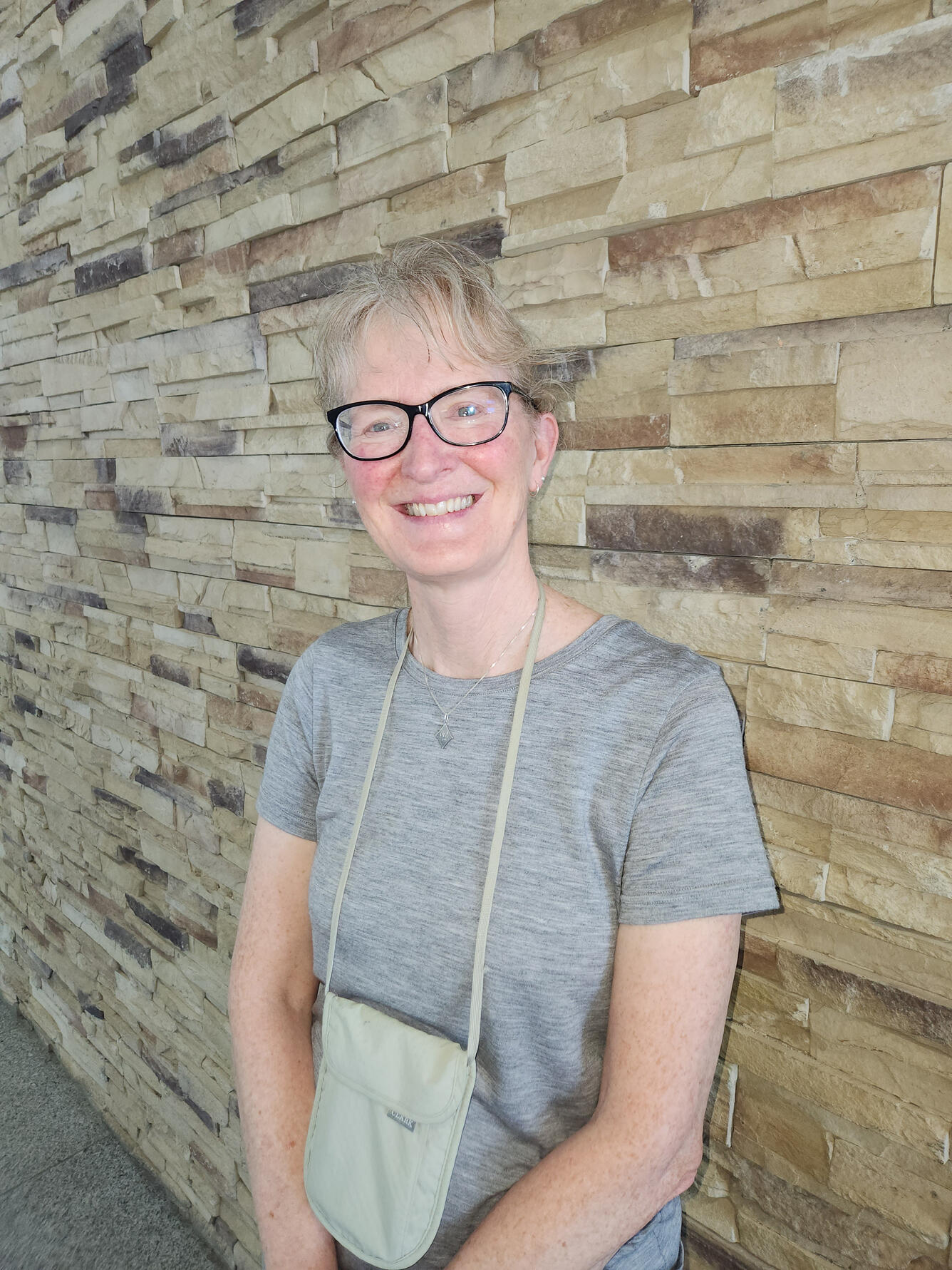 female scientist, smiling, wearing glasses, gray shirt, purse, standing near a rock wall.