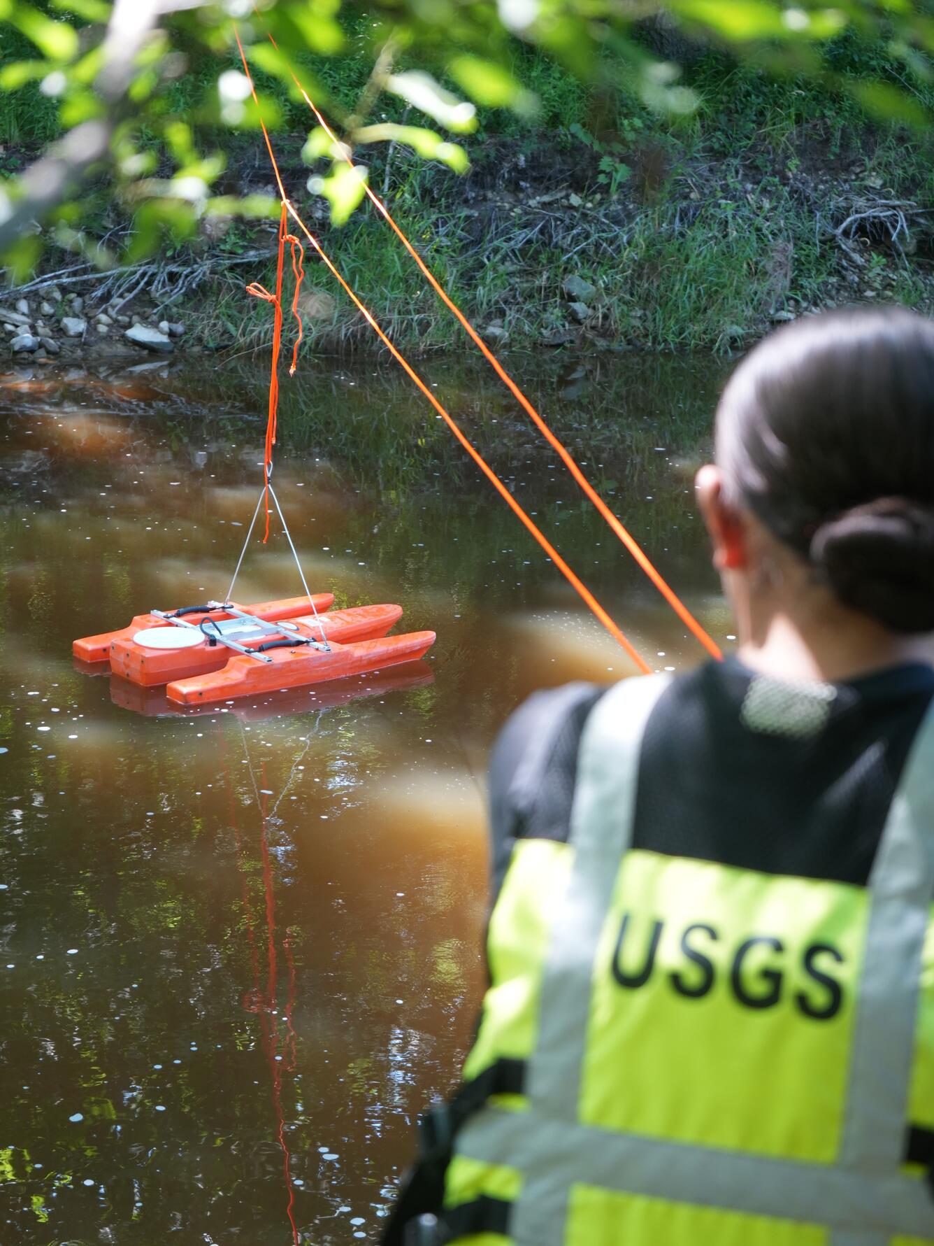 An acoustic Doppler current profiler is in focus with a person wearing a USGS personal flotation device in the foreground