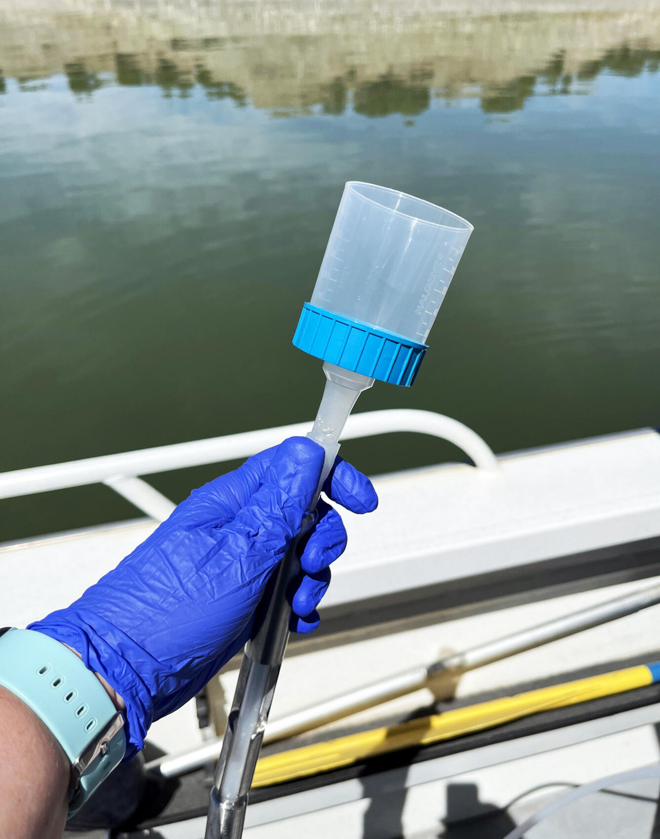A hand with a blue glove on holds an eDNA sampling filter next to the edge of a boat in a reservoir