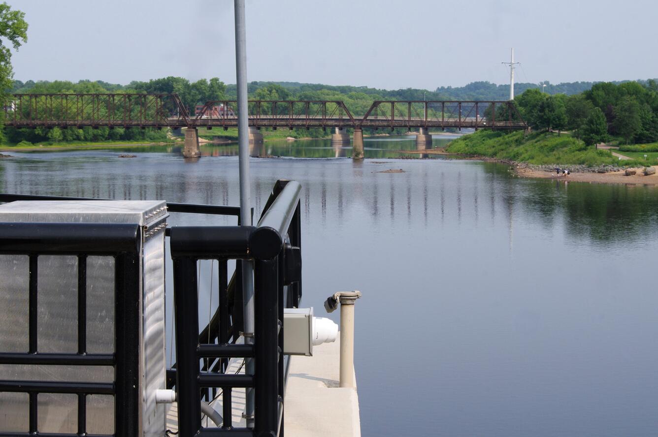 A white web camera looks over a river with a railroad bridge in background on a summer day