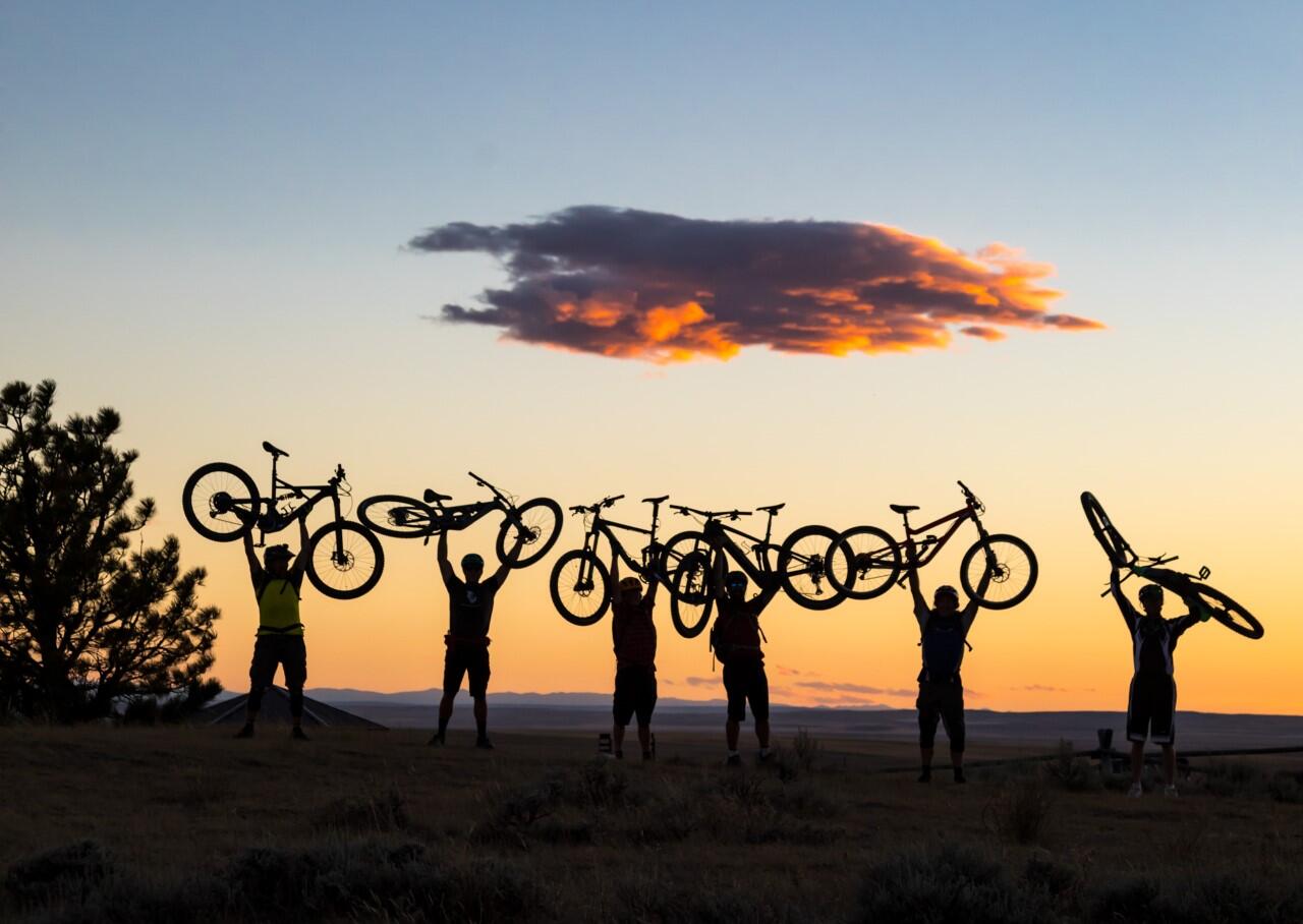 six people in shadow hold bikes over their heads, sunsetting in the background