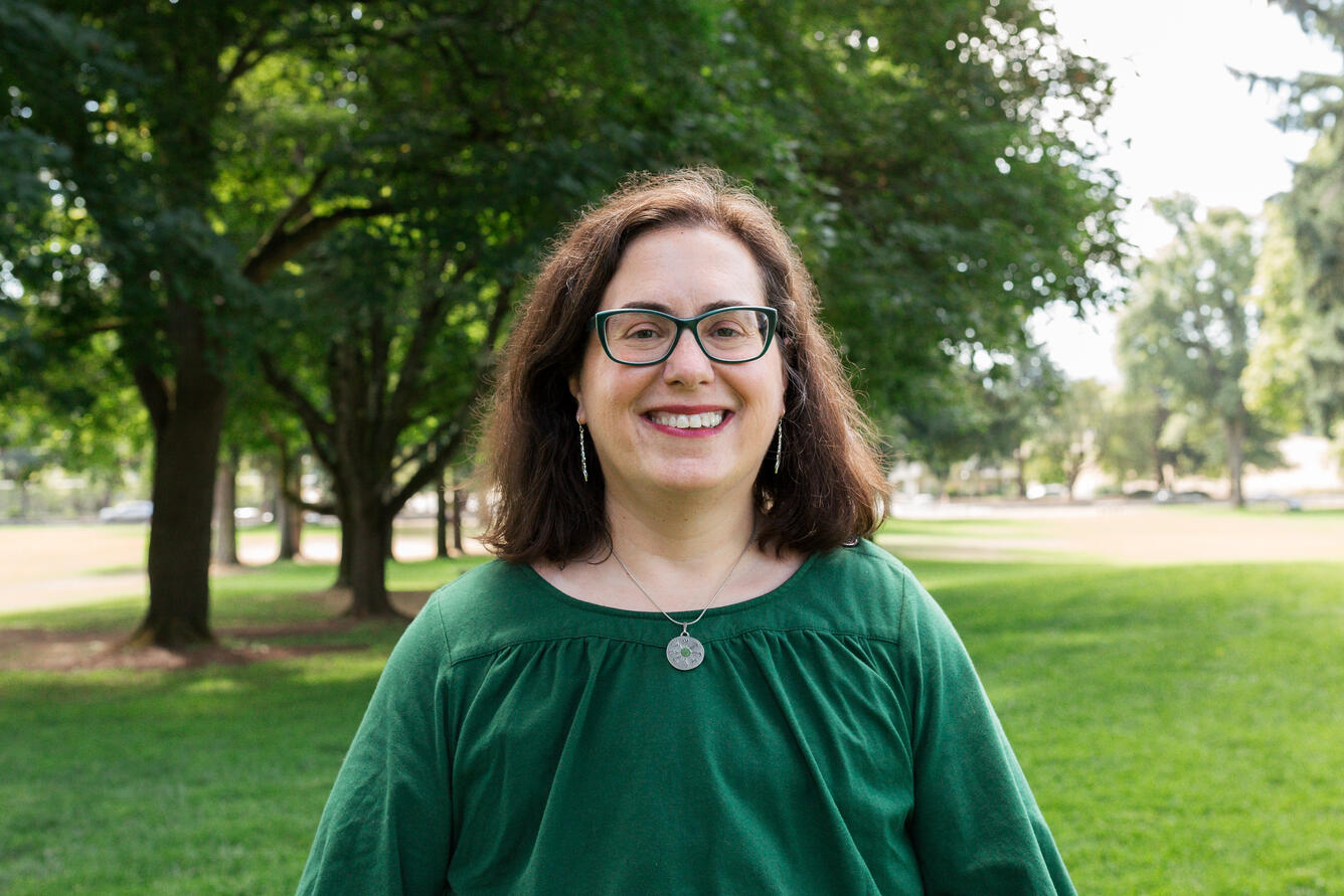 A woman with dark, shoulder length hair and a green shirt smiles for her picture while standing in a park.