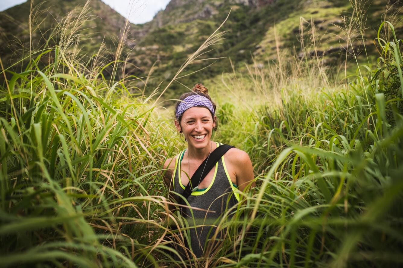 A woman standing in a tall grassy field with mountains behind her. She is smiling and wearing a grey tank top with a light purple bandana in her dark hair, which is pulled into a bun hairstyle.
