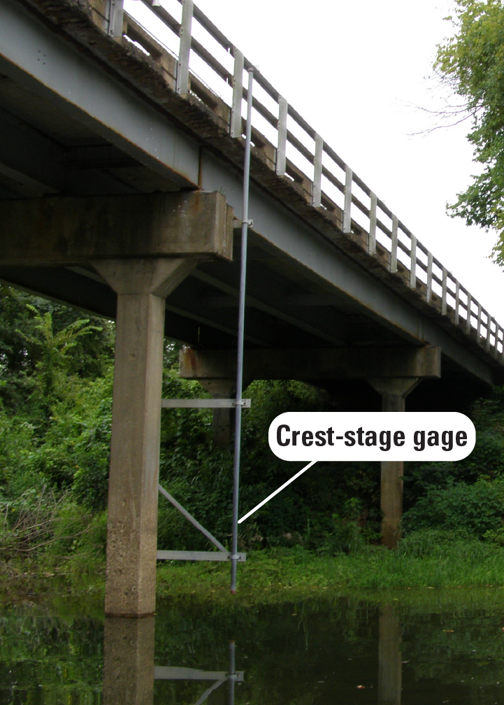 A long pipe is mounted to a bridge pier, stretching from the road railing down to the stream underneath.