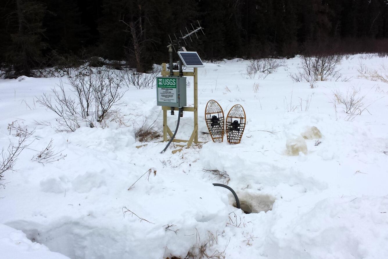 A pair of snowshoes rests in the snow next to a small streamgage mounted on a wooden frame, with antenna and solar panel.