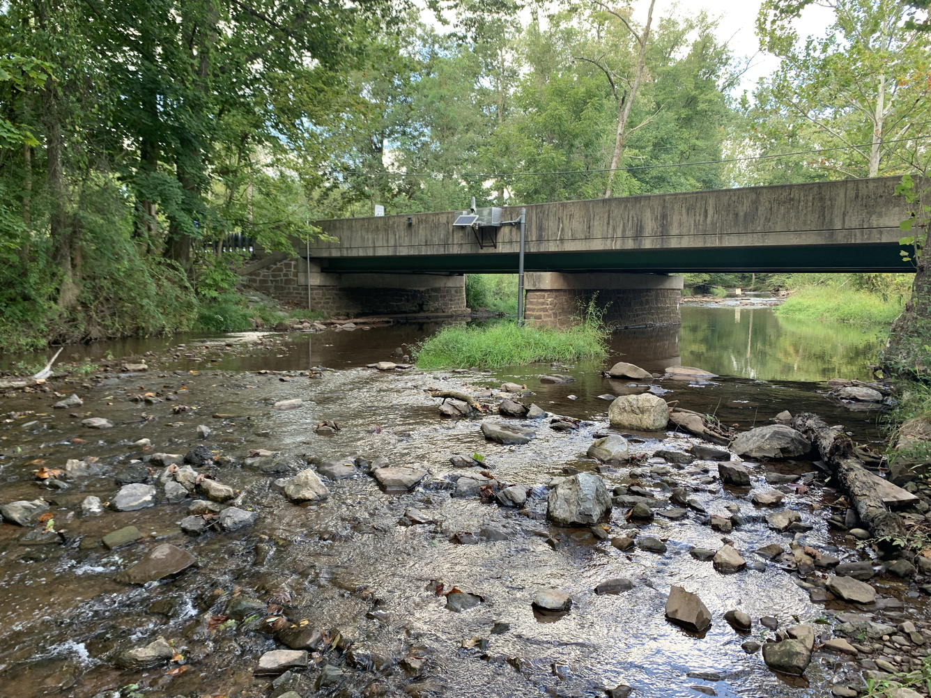 Facing upstream at concrete bridge with USGS gage box attached. Various sizes of rocks and low flow in stream channel. 