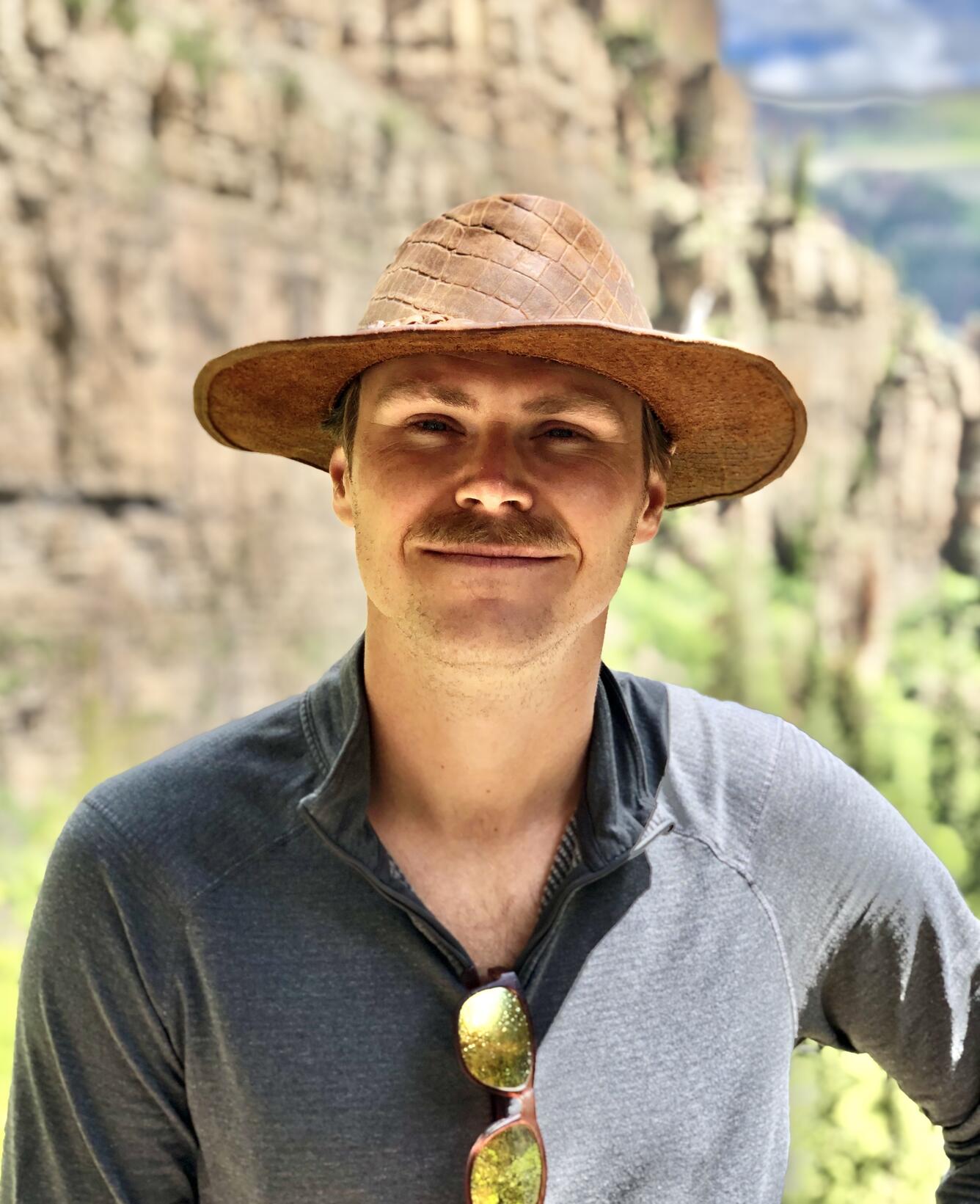 man wearing a tan wide brim hat, blue shirt, is standing near a rocky mountain